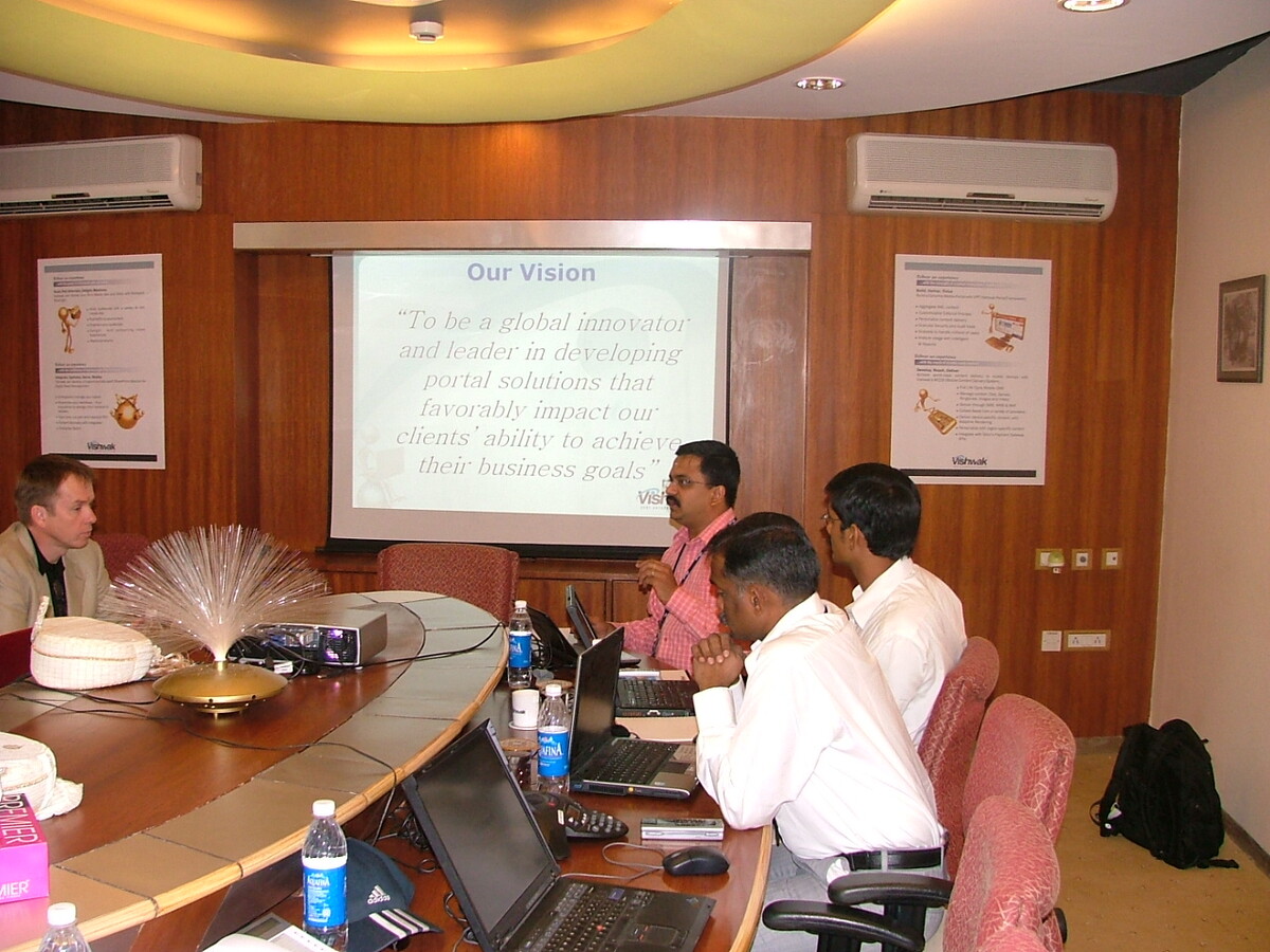 Team meeting in a wood-panelled conference room, colleagues seated around a table with laptops reviewing a projector slide titled 'Our Vision'